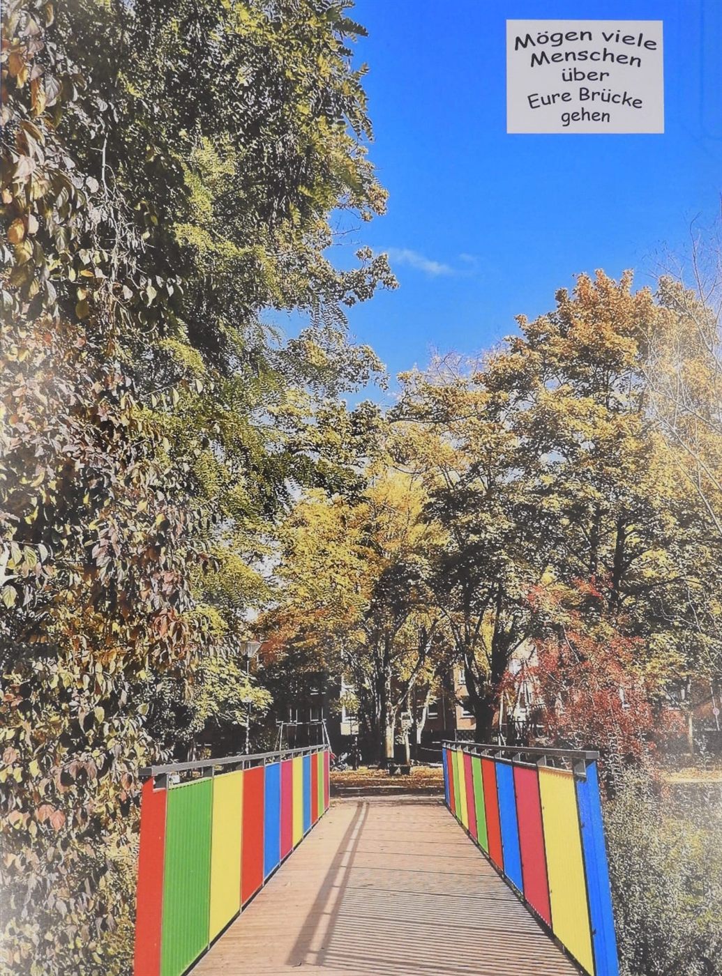 Bunte Fußgängerbrücke im Herbst, daran ein Schild mit der Aufschrift „Mögen viele Menschen über Eure Brücke gehen“.